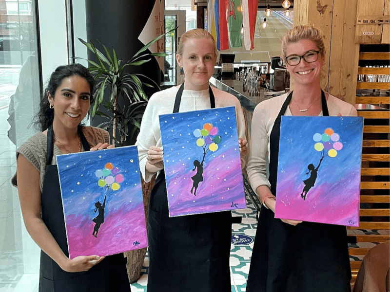 Three women hold painted canvases of a girl holding balloons at a Prime Pass painting class
